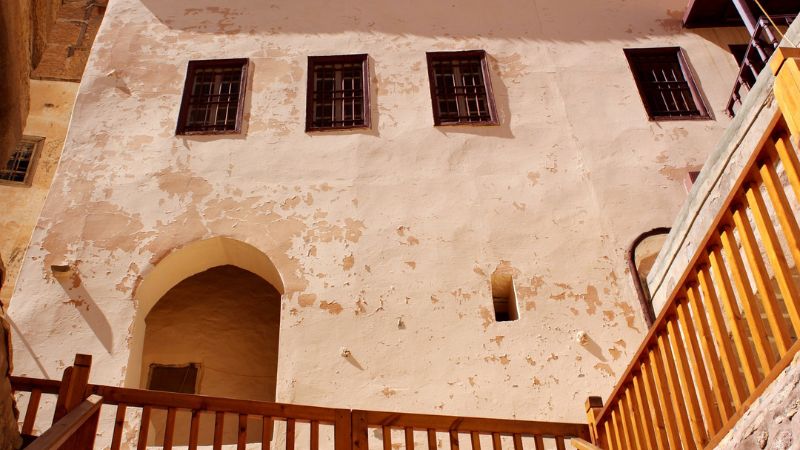 View of an aged building with chipped white walls, three arched windows, and a wooden balcony in an old monastery.