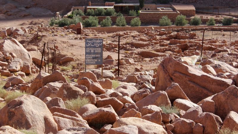 "A rugged landscape filled with reddish-brown rocks in the foreground with Saint Catherine's Monastery visible in the background, nestled at the base of a mountain."