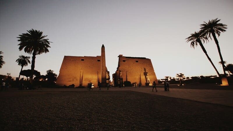 Luxor Temple at dusk, showing the illuminated temple walls, an obelisk, and palm trees against a twilight sky