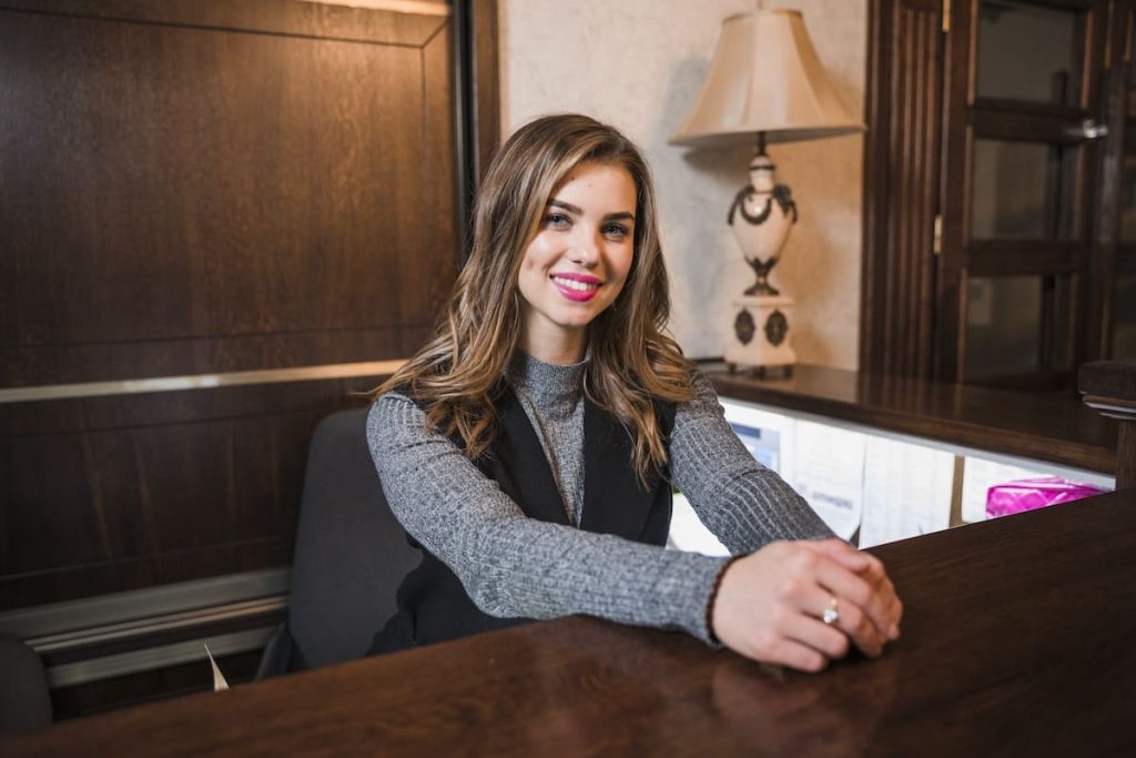 A cheerful young woman with long hair and bright lipstick, smiling warmly at a reception desk decorated with a classic lamp and wood paneling