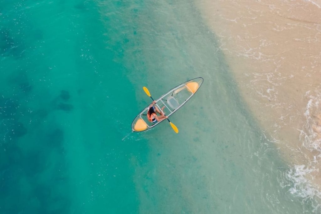 Overhead shot of a person in a yellow kayak paddling through vibrant turquoise waters close to a sandy beach.