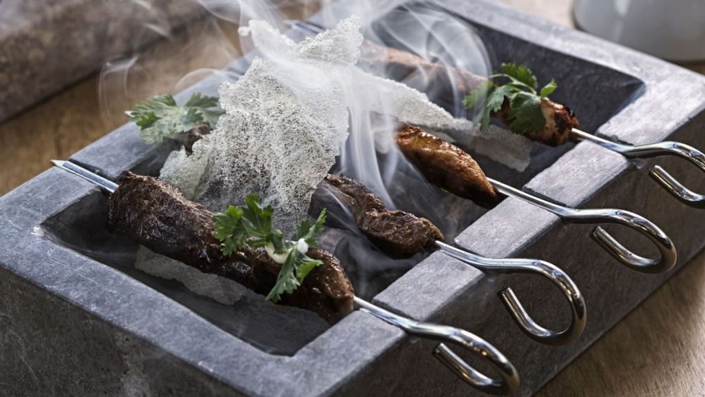 Grilled meat skewers on a gray stone slab, emitting steam, decorated with white, crisp garnish and green parsley