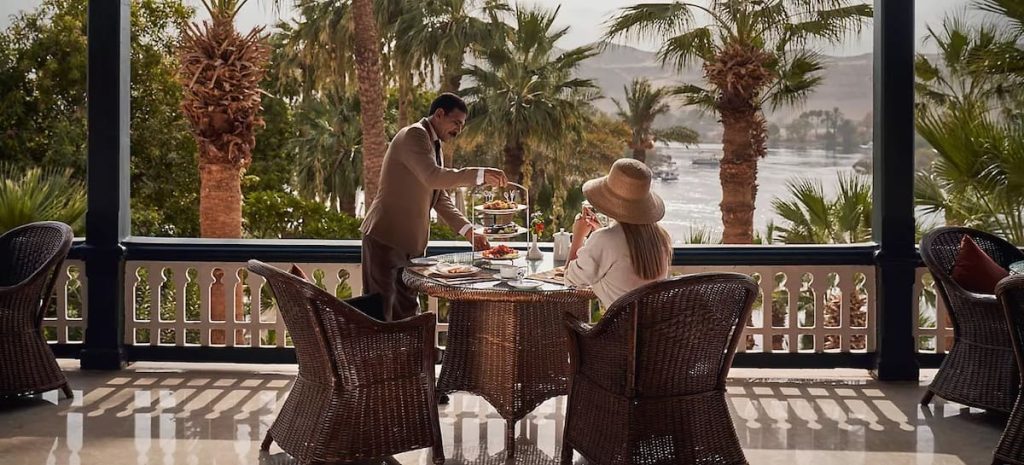 A woman in a hat seated at a dining table being served by a waiter, with lush palm trees and the Nile River in the background.