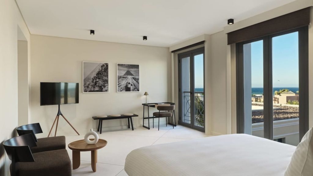 A stylishly furnished hotel room featuring a large bed, black contemporary chairs, and monochrome photos of pyramids on the wall, with a balcony overlooking the ocean