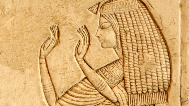 Bas-relief depiction of Queen Nefertiti in traditional Egyptian attire, with raised hands in a gesture of worship or blessing