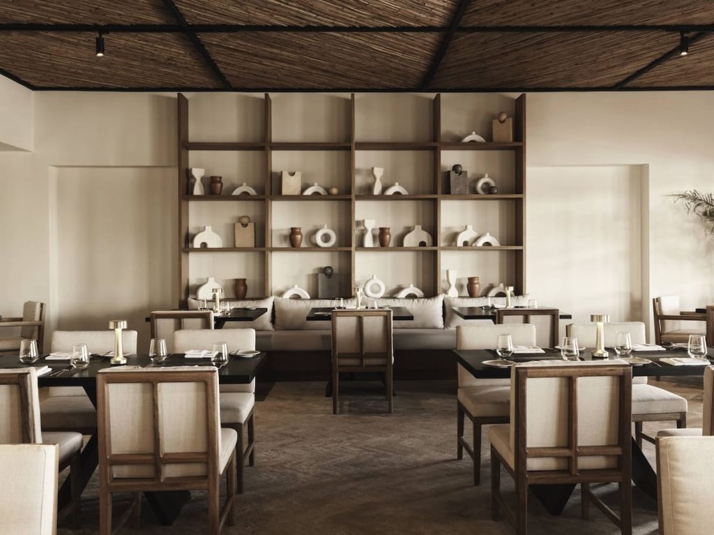 Interior of a sophisticated restaurant featuring large shelves with pottery, wooden tables, and plush seating under a thatched roof.