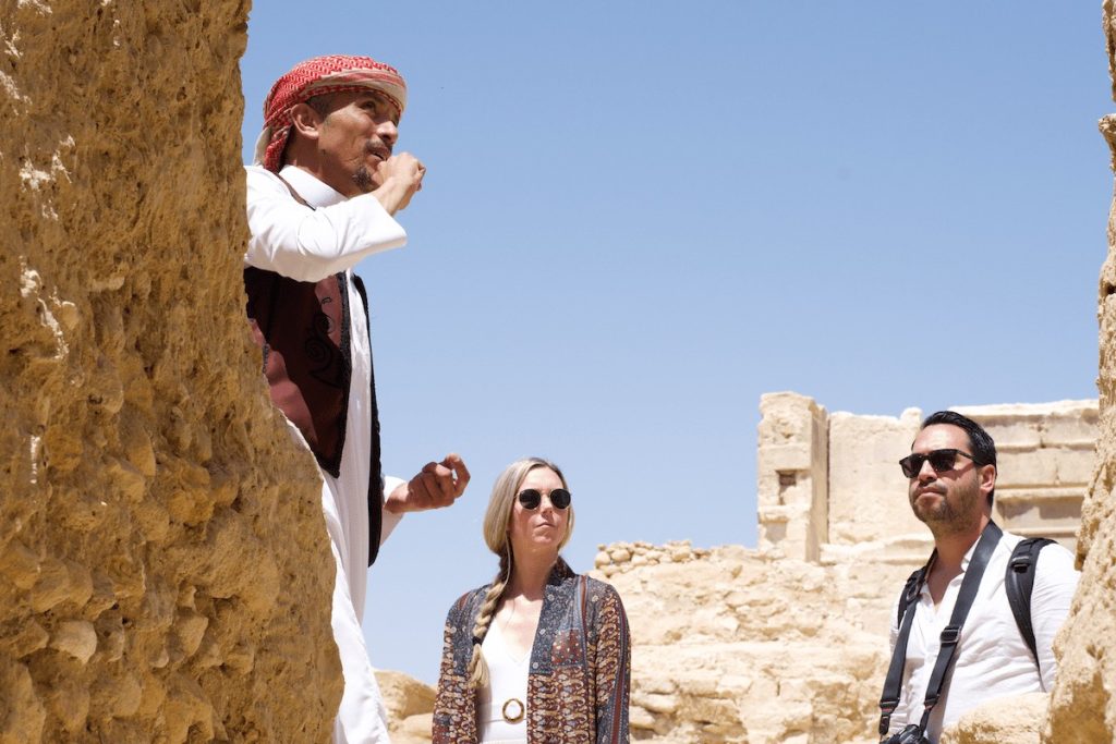 A tour guide in traditional attire explains historical details to two intrigued tourists at the Oracle Temple in Egypt."