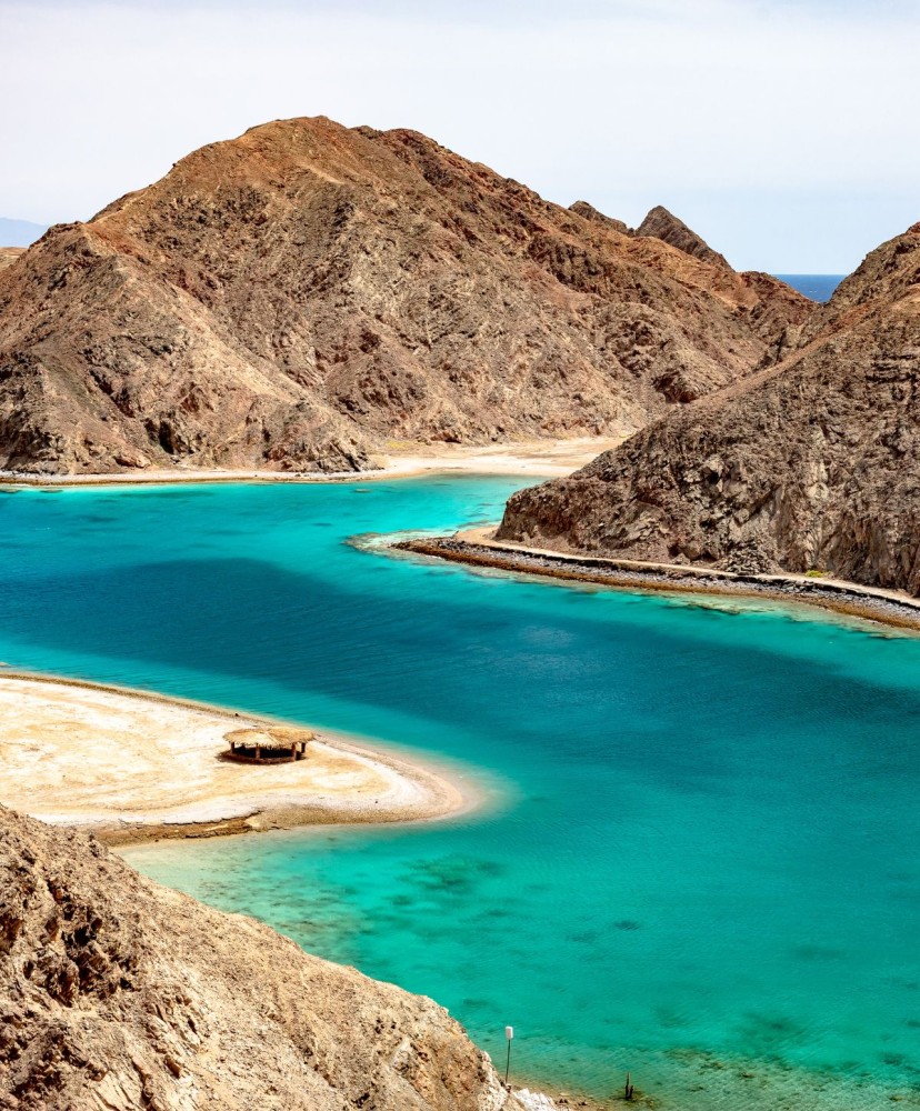 A scenic view of Taba's turquoise waters nestled between rugged Sinai mountains, with a sandbar and boat