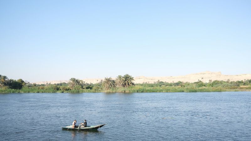 A small boat with two people floating on the tranquil waters of the Nile, surrounded by lush riverbanks.