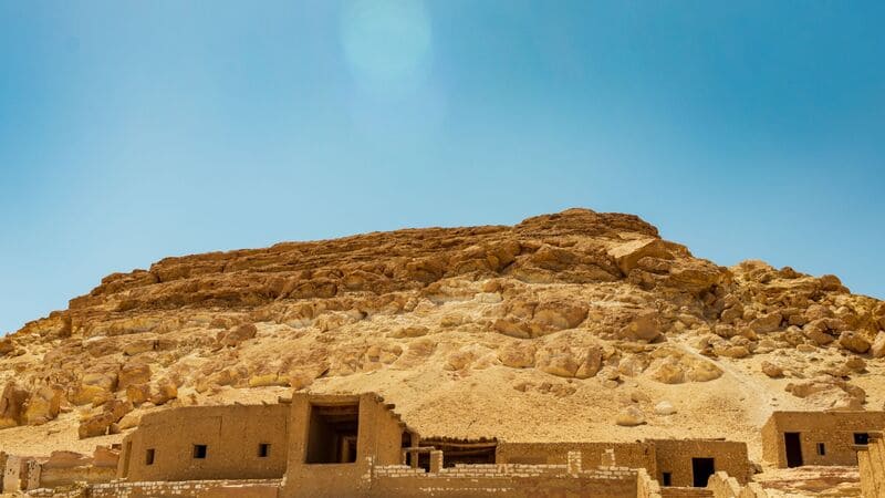 Ancient mud-brick structures at the base of a rocky hill under a clear blue sky