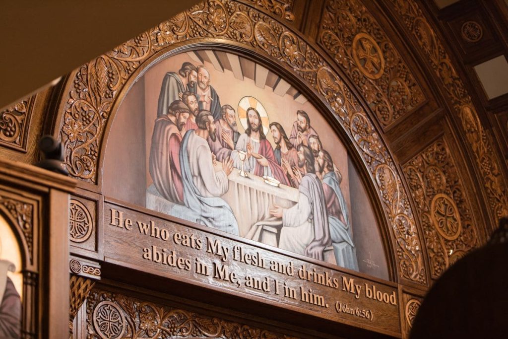 Carved wooden arch depicting The Last Supper, with Jesus and disciples, surrounded by ornate floral motifs and a biblical quote from John