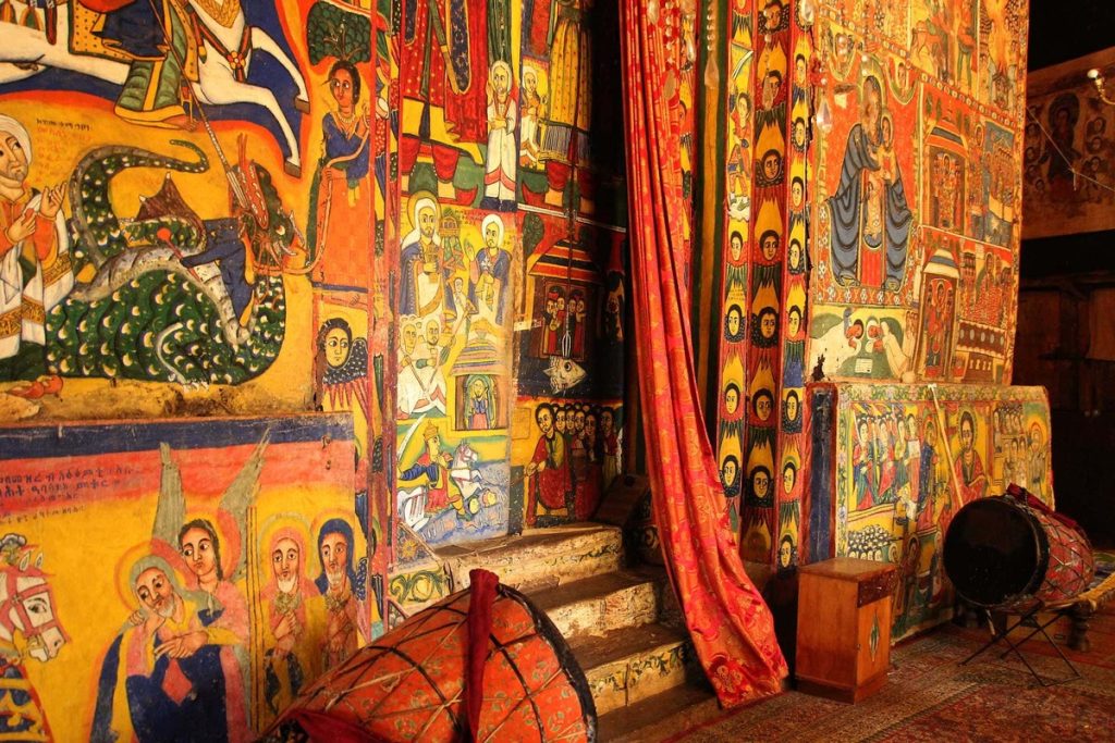 Colorful wall paintings in an Ethiopian church depicting various religious stories, including saints and biblical scenes, framed by ornate curtains.