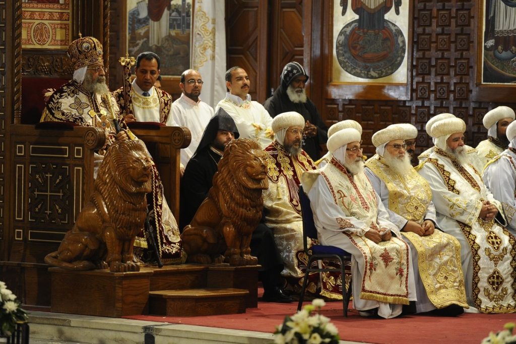 Coptic clergy in ornate vestments seated during a religious ceremony in a church adorned with intricate wood carvings and icons.