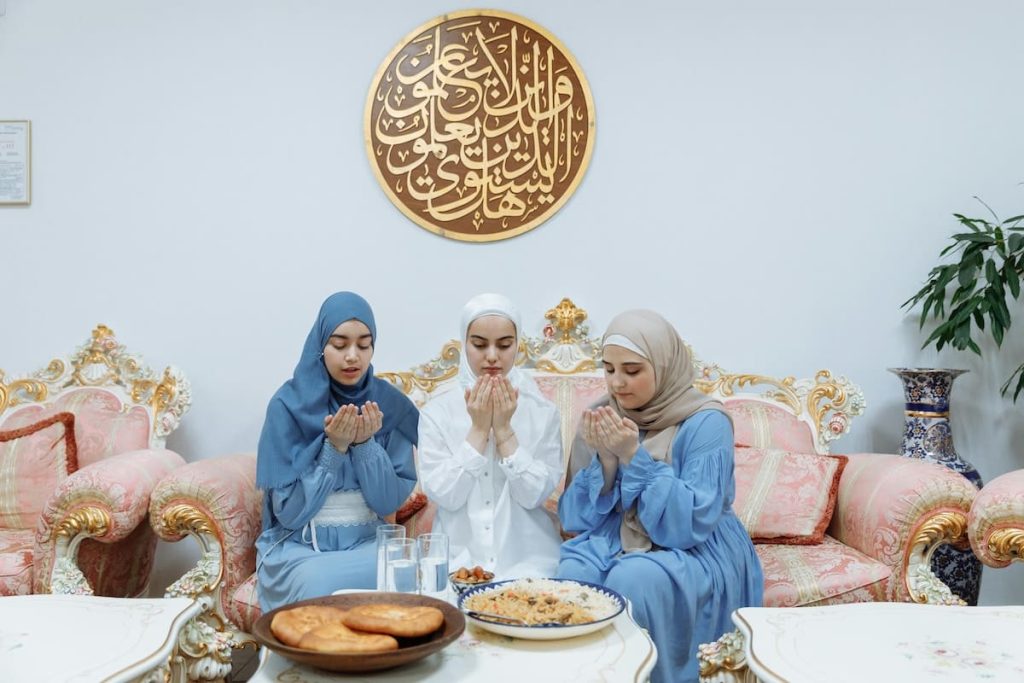 Three women in hijabs praying before eating, seated at a decorated table under Arabic calligraphy in a luxurious setting.