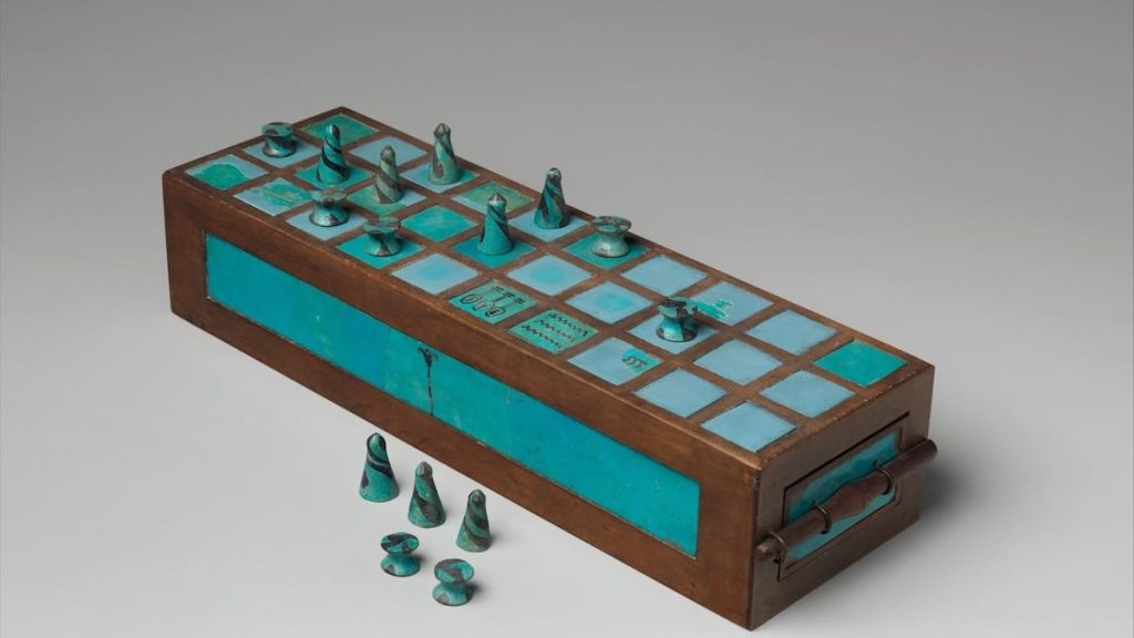 A photograph of an ancient Egyptian Senet game board with blue and white squares, bronze pieces, and a drawer.