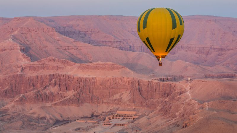 A green and yellow hot air balloon floats over the Valley of the Kings at dawn