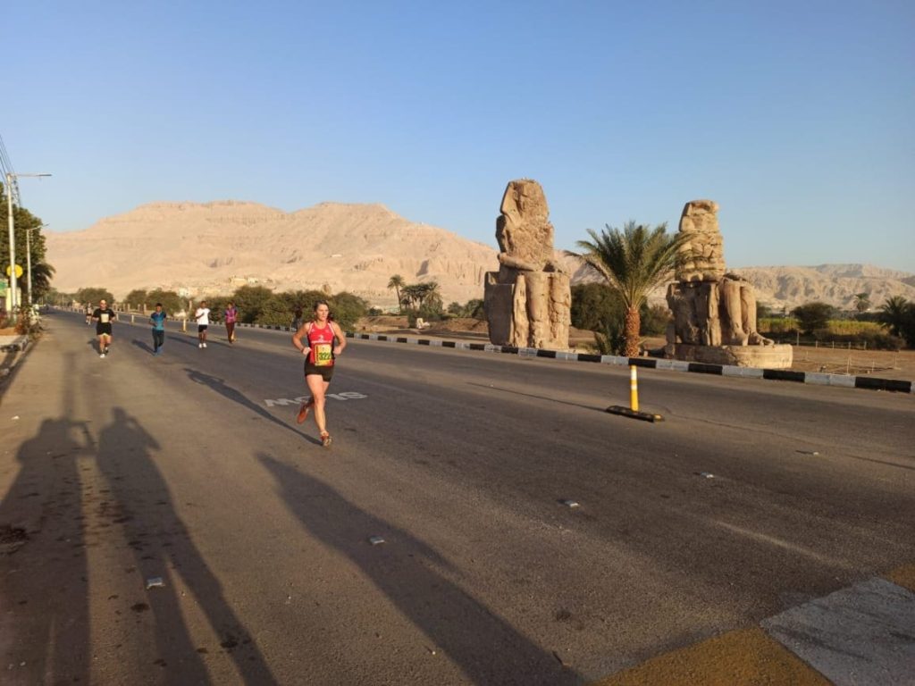 Runners participating in a marathon on a sunlit road, with the Colossi of Memnon and mountains in the background, Luxor, Egypt