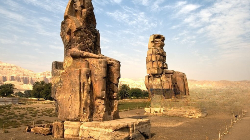 Two colossal statues known as the Colossi of Memnon under a partly cloudy sky.