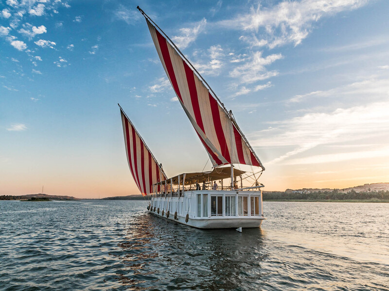 A traditional Egyptian dahabiya with red and white striped sails cruising on the Nile River at sunset