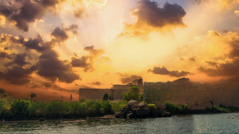 A dramatic sunset over an ancient temple on the Nile, with golden rays breaking through the clouds.