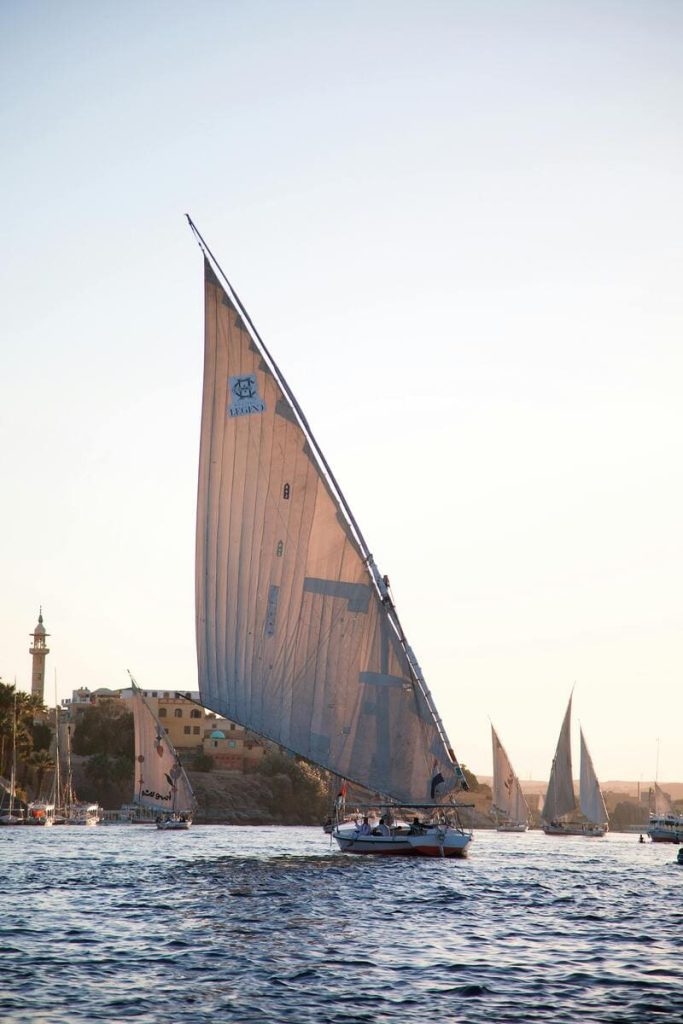 Traditional Egyptian felucca boats sailing on the Nile River at sunset with a minaret and buildings in the background.