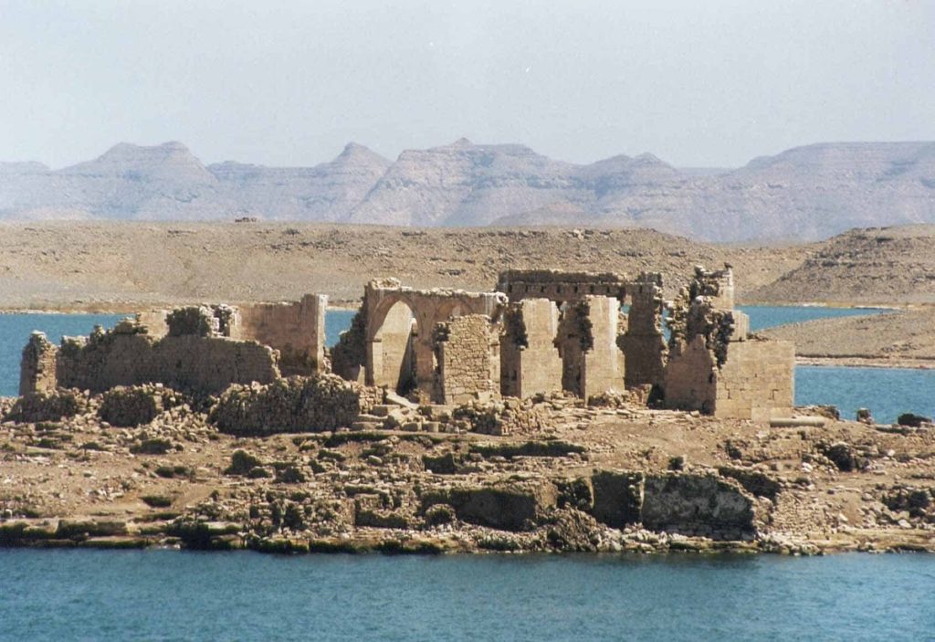 Ruins of Qasr Ibrim stand against a backdrop of rugged mountains and a blue lake, showcasing the remains of arches and stone walls in a remote desert landscape