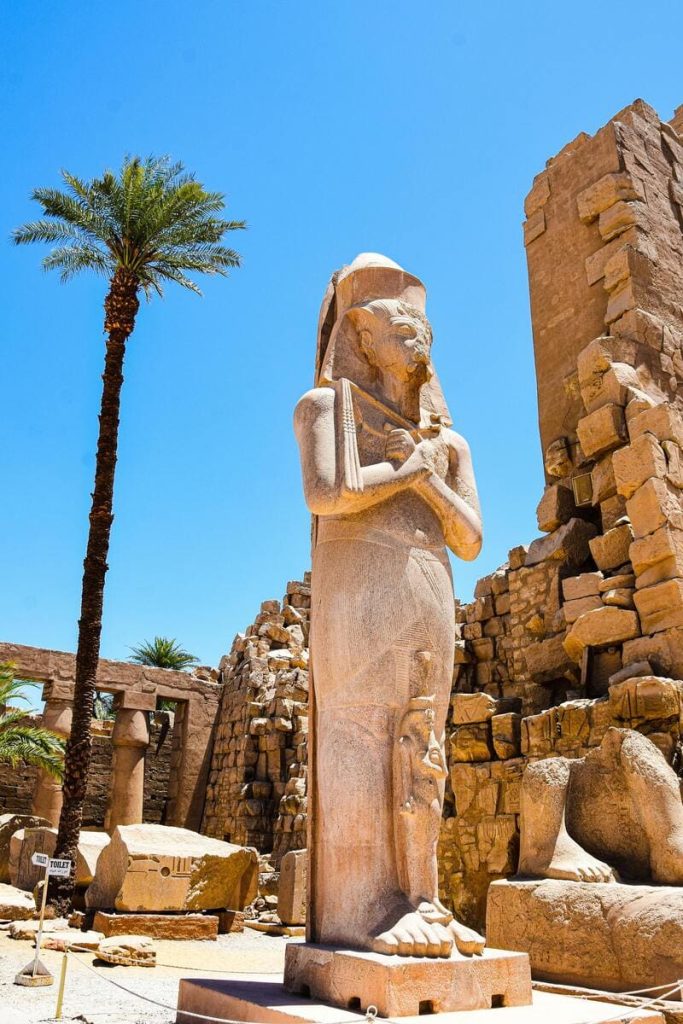 Ancient statue of Pharaoh Ramses II at the Temple of Karnak with palm trees and ruins in the background under a clear blue sky