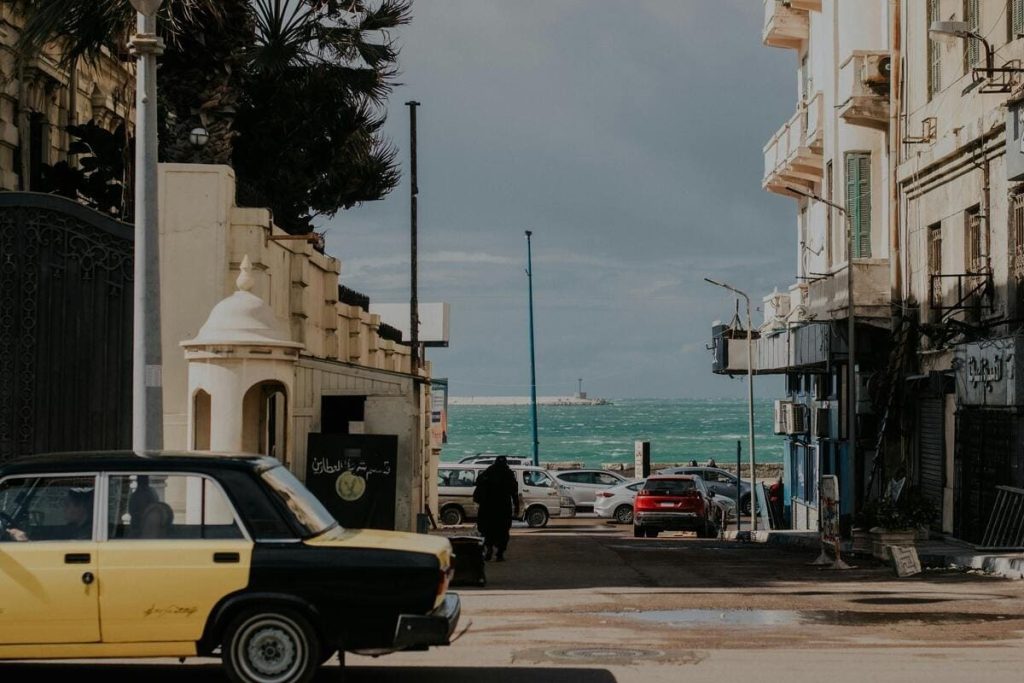 A street in Alexandria with a yellow and black taxi, buildings, and a view of the sea.