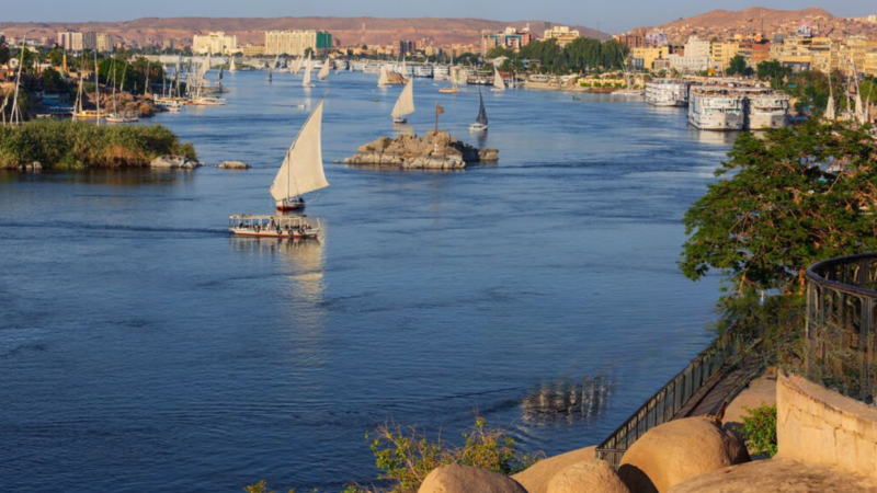 A serene view of the Nile River with sailboats and the cityscape of Aswan in the background.