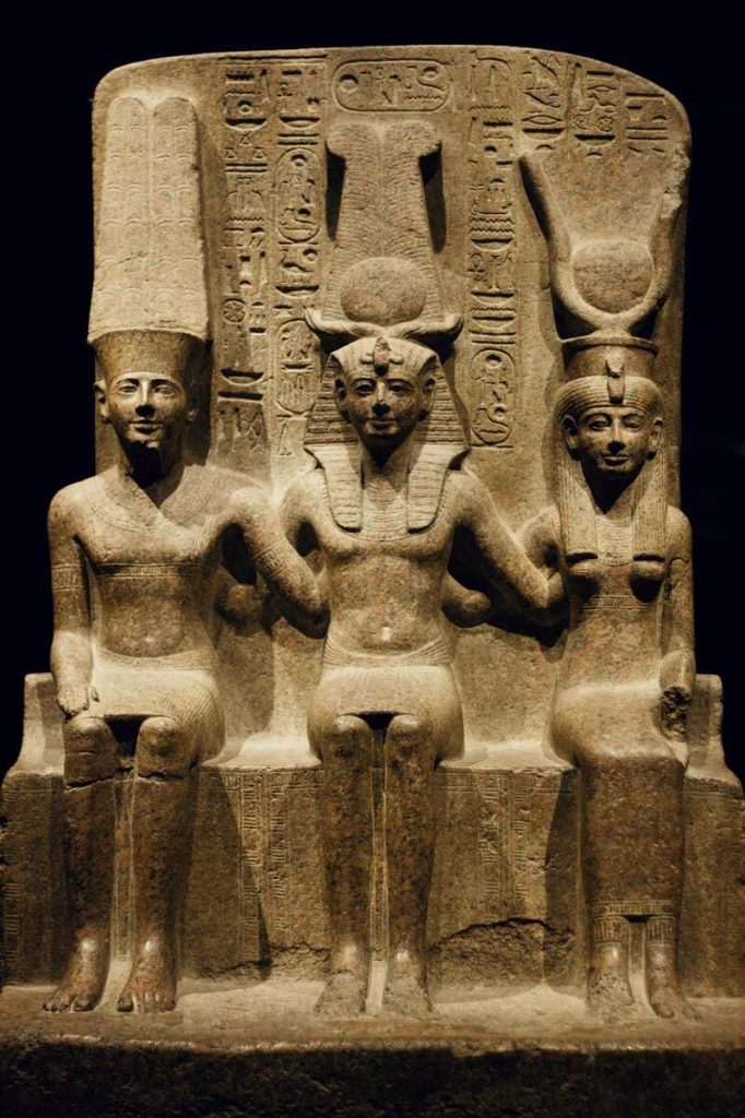 Ancient Egyptian statue depicting a pharaoh and two goddesses with hieroglyphics in the background