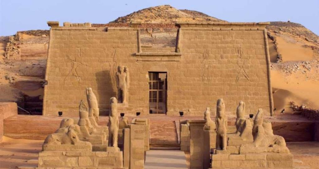 A front view of the Wadi el-Seboua temple in Egypt, flanked by statues of sphinxes leading to the entrance, set against a backdrop of desert hills.