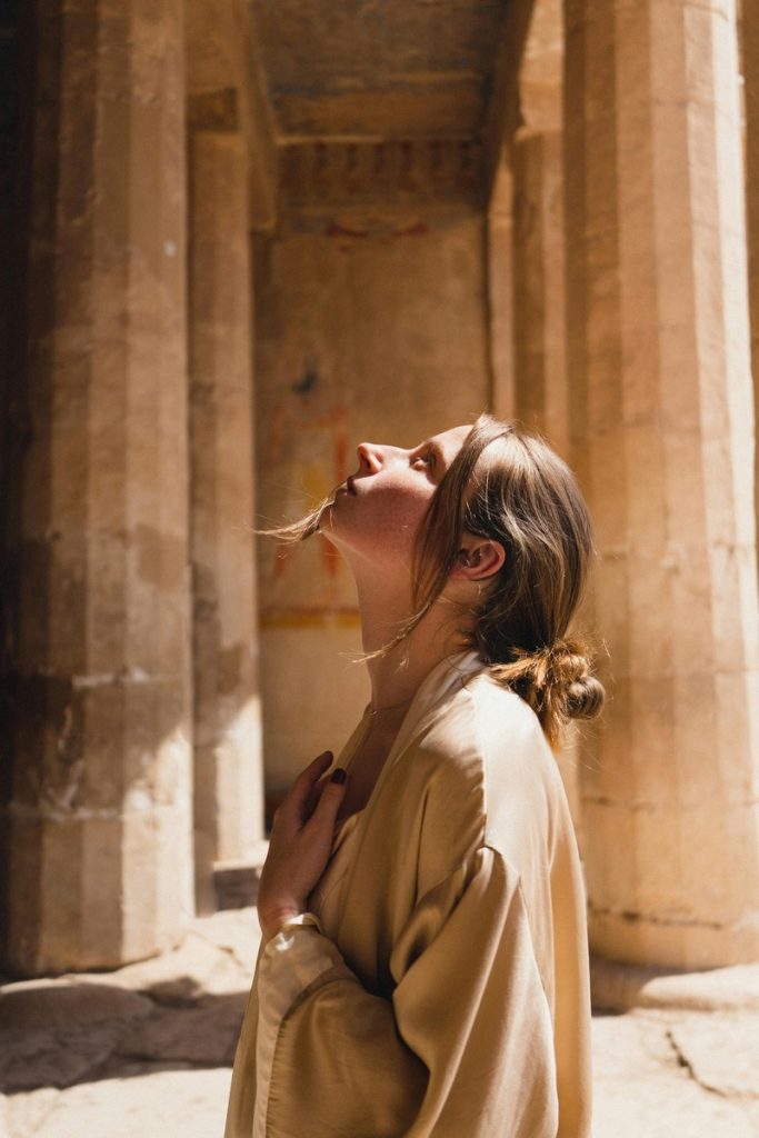 "A woman stands inside an ancient Egyptian temple, gazing upwards with reverence."