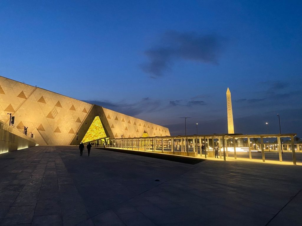 A modern architectural view of the Grand Egyptian Museum at twilight, featuring its geometric facade and an ancient obelisk, illuminated under a fading blue sky.