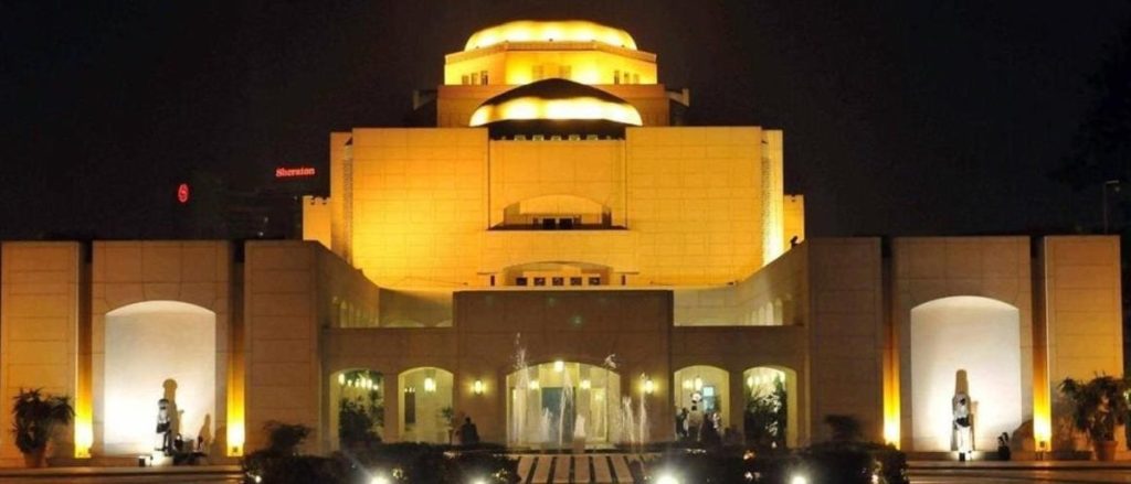 Nighttime view of the Cairo Opera House, illuminated in warm yellow lights, showcasing its modern architecture with a central dome and open gateways