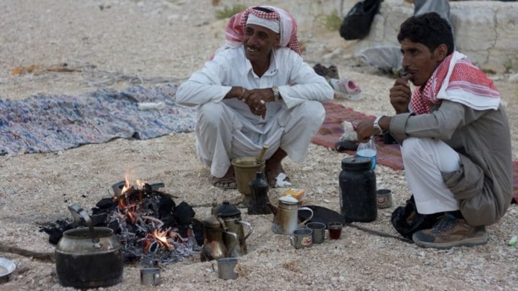 Two men in traditional Bedouin attire sitting on a rug in the Sinai desert, enjoying tea by a small fire with teapots and cups.