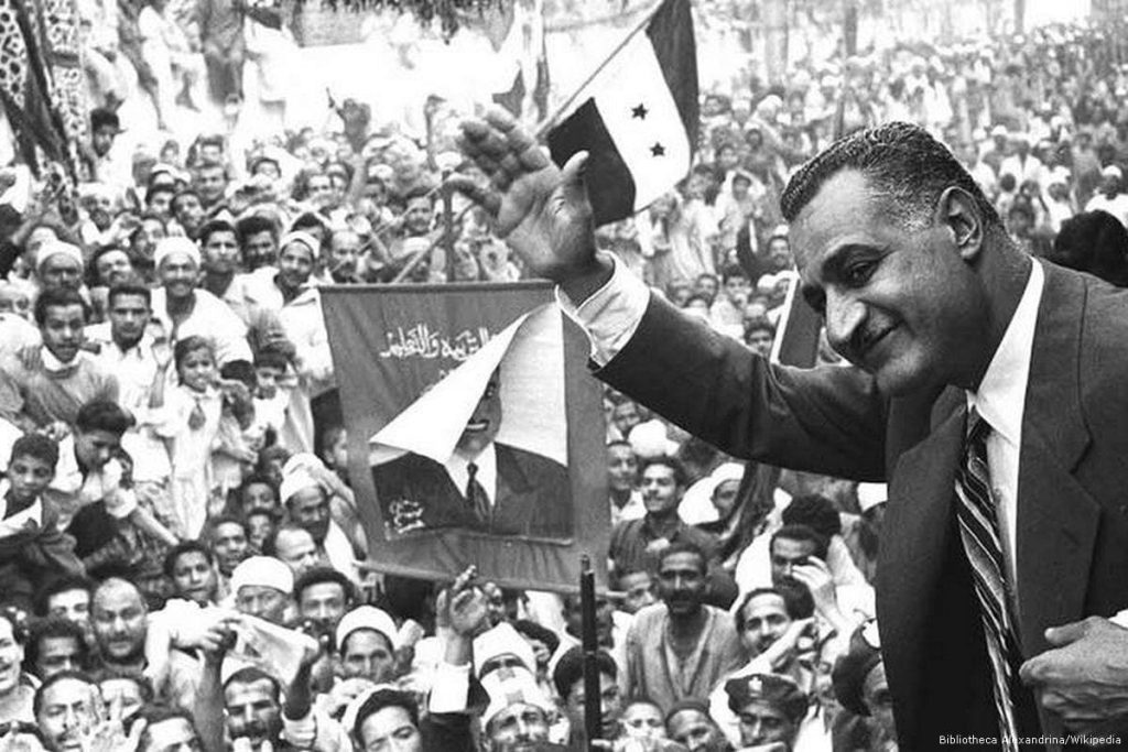 Black and white photograph of Egyptian President Gamal Abdel Nasser waving to a large crowd in Mansoura, Egypt, in 1960. He is surrounded by joyful citizens holding flags.