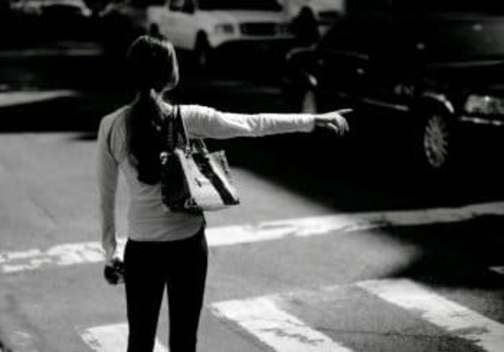 Black and white image of a woman hailing a taxi on a busy city street. She's wearing a long sleeve shirt and jeans, carrying a camera and a shoulder bag.