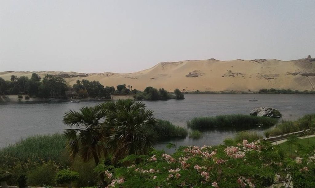 Scenic view of the Nile River in Egypt, featuring lush greenery, a vast desert dune in the background, and a small boat floating peacefully on the water.