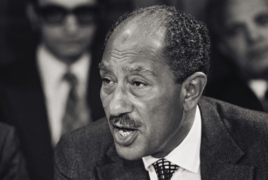 Black and white portrait of Anwar Sadat, former President of Egypt, speaking at a conference. He appears in a suit with a distinctive tie, surrounded by other attendees.