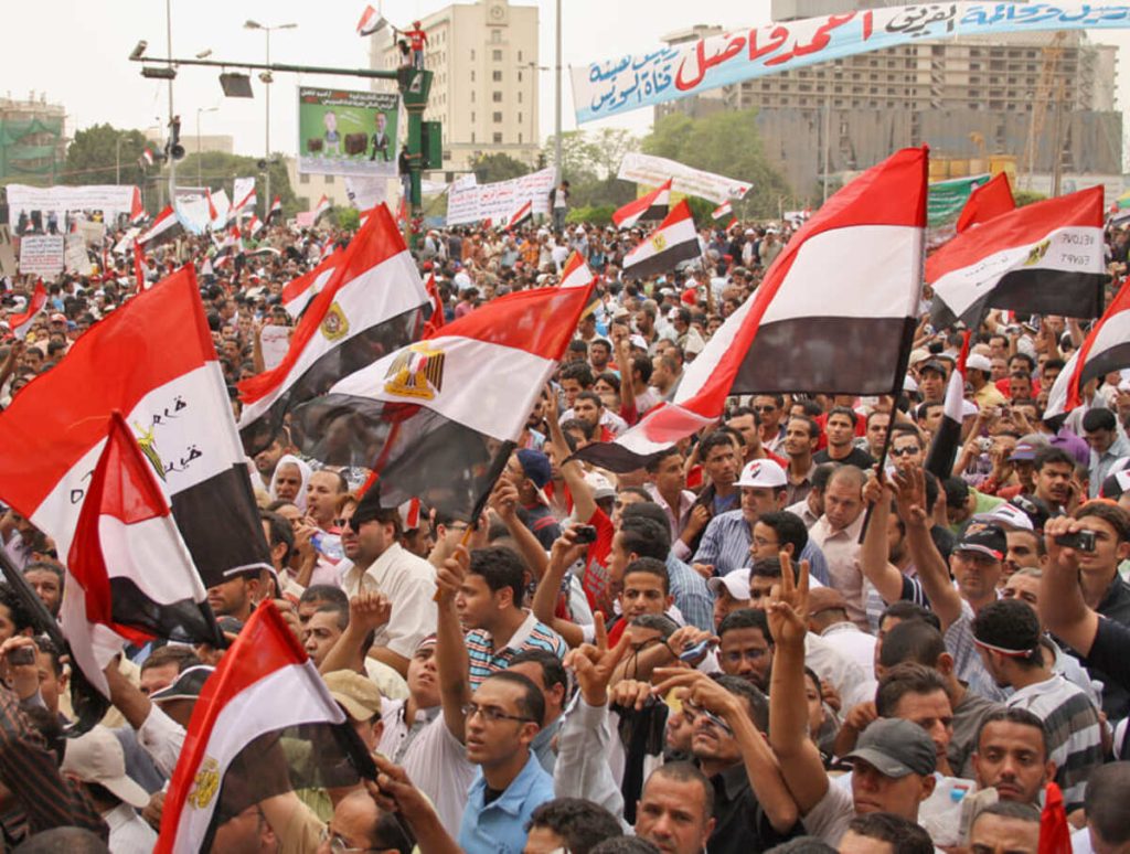 A dense crowd of protesters waving Egyptian flags in Tahrir Square, Cairo, with banners and placards, expressing political demands.