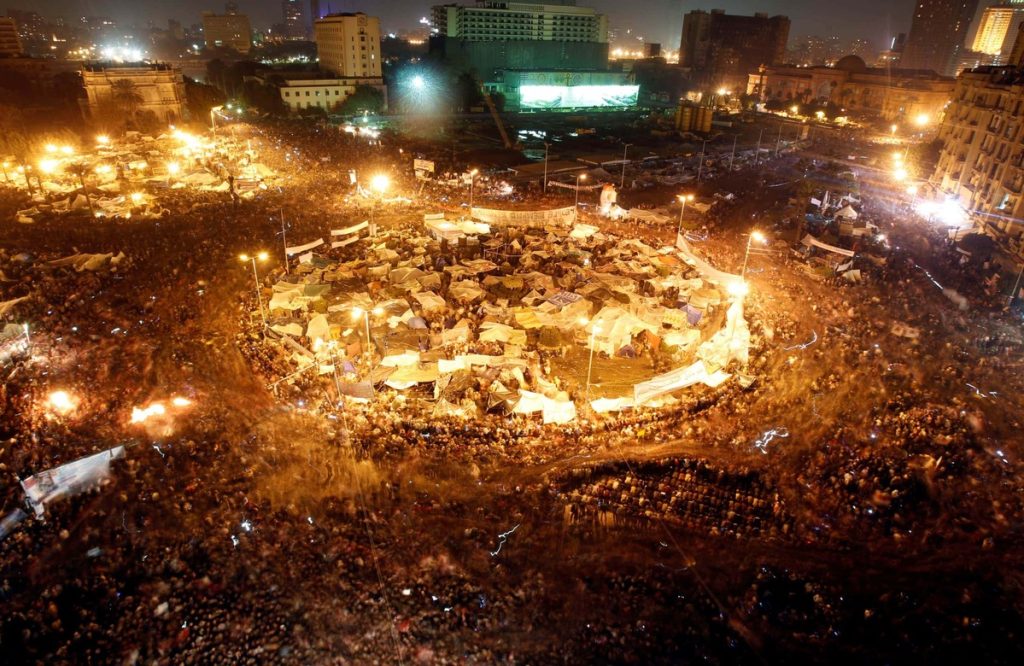 Nighttime aerial view of Tahrir Square in Cairo, Egypt, during a major protest. The square is densely packed with thousands of protesters, tents, and bright lights.