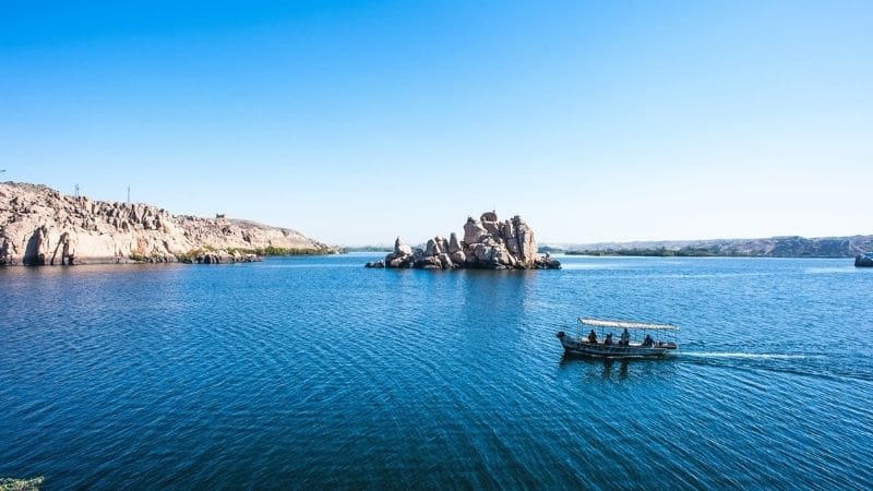 : A small boat glides across the calm, blue waters of Lake Nasser under a clear sky with rocky islands in the distance.