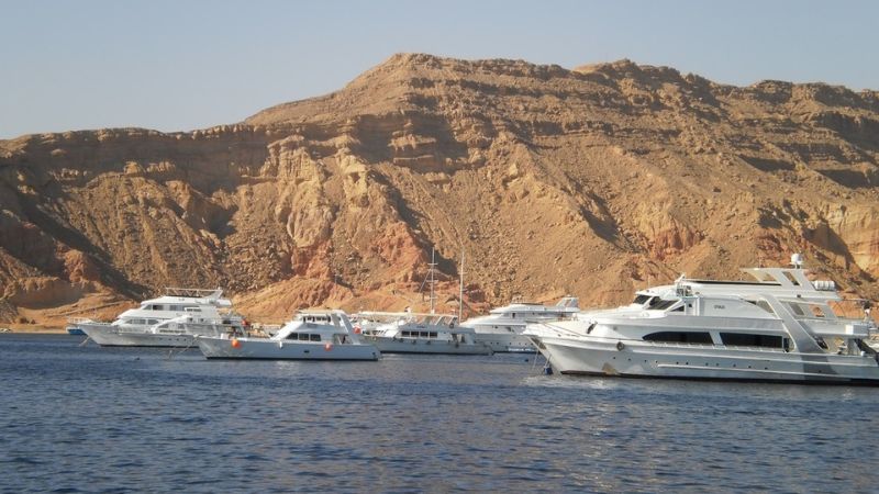 White yachts anchored near the rugged coastline of the Gulf of Aqaba, surrounded by desert cliffs.