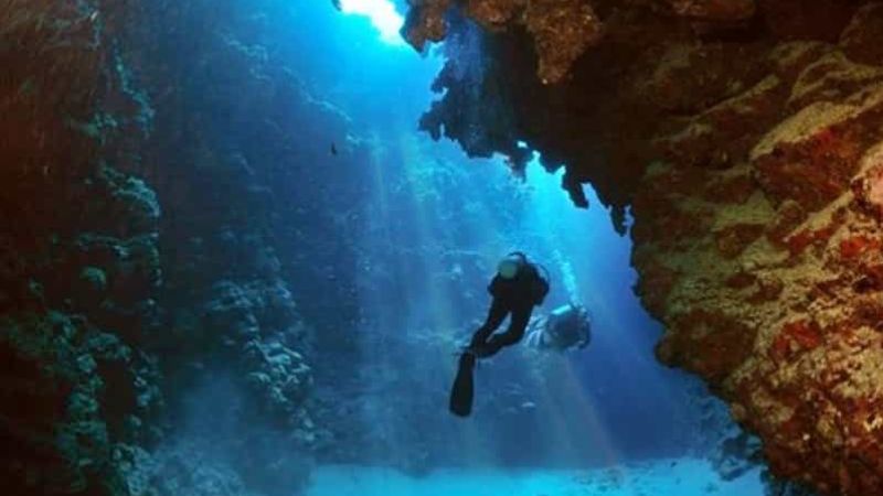 Scuba diver exploring an underwater cave surrounded by vibrant coral walls, with sunlight streaming through the clear blue waters in the Red Sea, Egypt