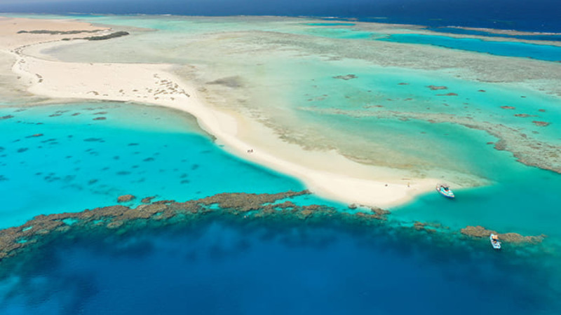 A stunning aerial view of a white sandy island surrounded by crystal-clear turquoise waters and coral reefs in the Red Sea, with two boats anchored nearby.