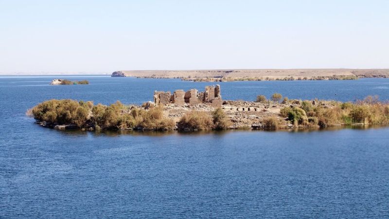 Ruins of an ancient settlement on a small island in a vast blue lake, surrounded by sparse vegetation and distant desert landscapes