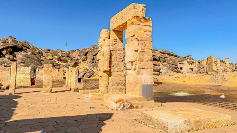 Ancient Egyptian temple ruins with sandstone columns and intricate carvings, surrounded by rocky desert terrain under a clear blue sky