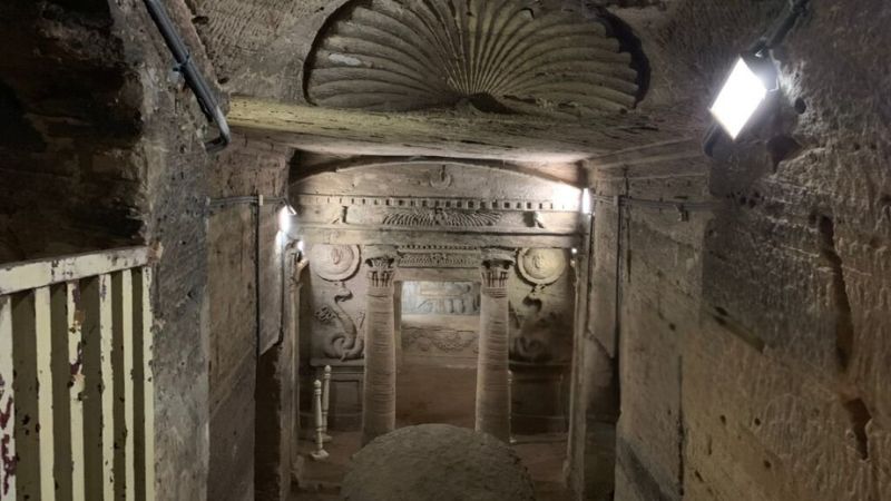 Ancient underground chamber in the Catacombs of Kom El Shuqafa, featuring carved arches and reliefs illuminated by lights