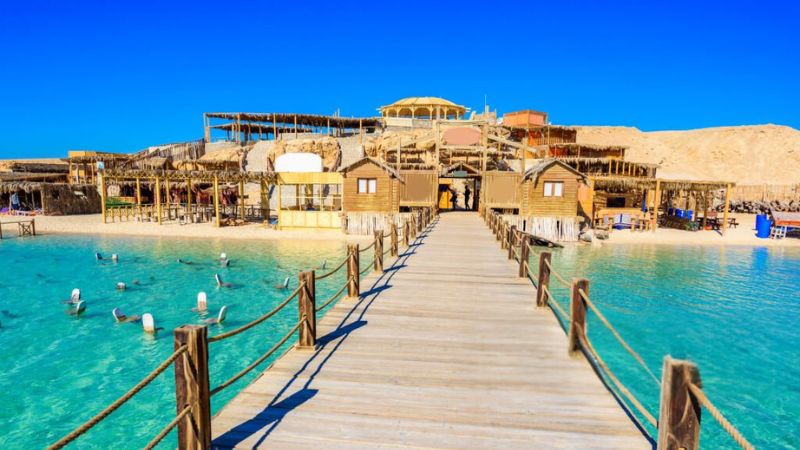 Wooden walkway leading to a vibrant beach resort with rustic huts, surrounded by turquoise waters and clear blue skies