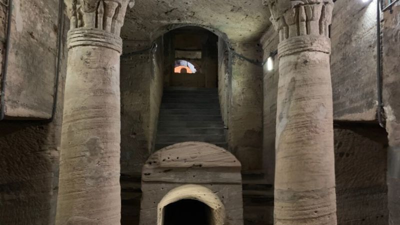 A pillared hallway within the Catacombs of Kom El Shuqafa, illuminated by soft lights and steeped in ancient architecture